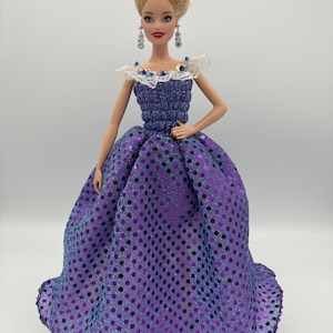 May include: A doll wearing a purple gown with a full skirt and a bodice adorned with sequins. The doll has blonde hair styled in an updo, dangling earrings, and a white lace collar.