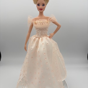 May include: A doll wearing a light peach-colored gown with a fitted bodice and a full skirt. The dress features delicate, sheer sleeves and is adorned with small, raised dots. The doll has blonde hair styled in an updo and is posed with one hand on her hip.