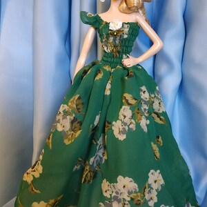 May include: A doll wearing a green floral dress with a white flower accent. The dress has a gathered waist and a one-shoulder design.