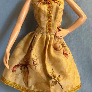 May include: A doll wearing a strapless, knee-length dress with a floral pattern in shades of yellow, brown, and red. The dress is trimmed with gold and adorned with orange floral accents. The doll is also wearing pink heels and dangling earrings.