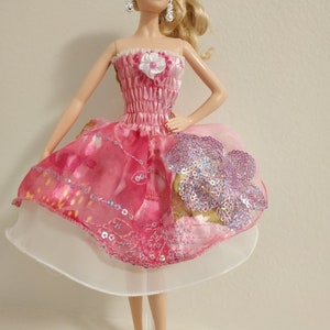 May include: A pink and white dress with sequins and a flower detail. The dress is on a doll with blonde hair and red shoes.