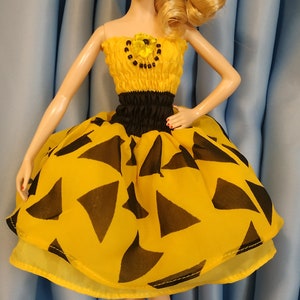 May include: A doll wearing a yellow dress with black triangle patterns. The dress has a black waistband and a flower detail. The doll has blonde hair, dangling earrings, and red heels. The doll is standing on a white stand.