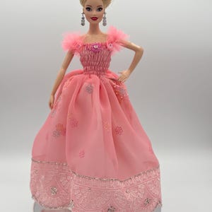 May include: A doll in a pink gown with a ruched bodice and a full skirt. The dress features delicate floral embroidery and a scalloped hemline. The doll has blonde hair styled in an updo, dangling earrings, and is posed on a reflective surface.