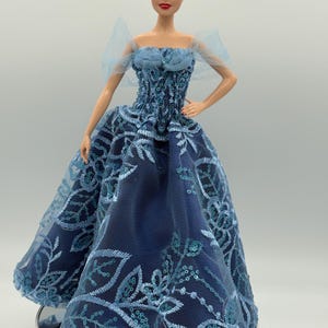 May include: A doll wearing a long, navy blue gown with intricate light blue floral embroidery and sequins. The doll has blonde hair styled in an updo and is posed with one hand on her hip. The gown features a sheer overlay.