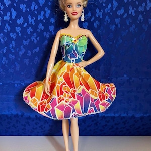 May include: A fashion doll wearing a strapless dress with a colorful geometric pattern in shades of red, orange, yellow, pink, and blue. The doll has blonde hair, dangling earrings, and pink high-heeled shoes.