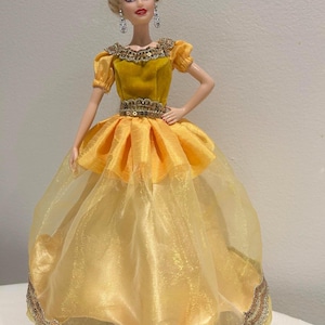 May include: A doll wearing a yellow gown with a layered skirt and a bodice adorned with sequins. The doll has blonde hair styled in an updo and is wearing dangling earrings. The dress features a sheer, flowing overskirt.