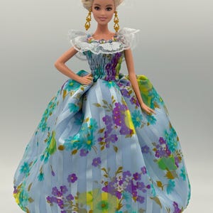 May include: A doll with blonde hair styled in an updo, wearing gold earrings and a light blue floral gown. The gown features a ruffled neckline and a full skirt with vertical stripes and floral patterns in purple, green, and yellow.