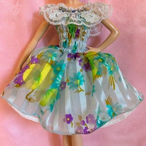 May include: A doll wearing a floral print dress with a white lace collar and matching earrings. The dress features a blue, green, and purple floral pattern with white vertical stripes. The doll is wearing pink high heels.