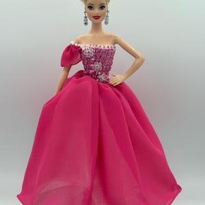 May include: A doll in a bright pink gown with a fitted bodice adorned with white floral accents and a single off-the-shoulder sleeve. The doll has blonde hair styled in an updo, dangling earrings, and is posed on a reflective surface.