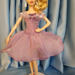 May include: A blonde doll wearing a lavender dress with floral print and red shoes. The doll is standing on a white pedestal against a blue background.