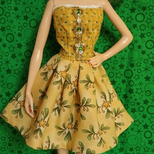 May include: A blonde doll wearing a yellow dress with green leaves and gold stars. The dress has a strapless bodice with green gem embellishments. The doll is wearing red shoes and has her hair styled in a braid.