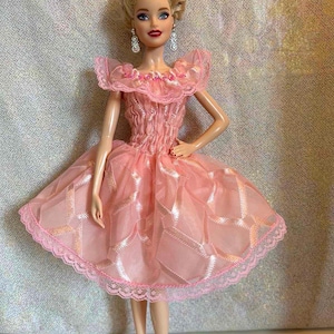 May include: A doll wearing a pink dress with a layered skirt and lace trim. The dress has a ruffled neckline and is paired with pink heels. The doll has blonde hair and is wearing dangling earrings.