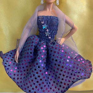 May include: A doll wearing a sparkly, strapless, knee-length dress with a polka dot pattern in shades of purple and pink. The doll has blonde hair, red lipstick, and dangling earrings. A sheer, lavender shawl is draped over the doll's shoulders.