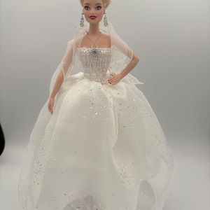 May include: A doll in a white wedding dress with a sparkling bodice and a full, layered skirt. The doll has blonde hair styled in an updo, and is wearing a veil, earrings, and a necklace. The dress is embellished with silver glitter.