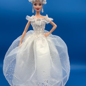 May include: A doll wearing a white wedding dress with a sheer, sparkling overlay. The doll has blonde hair, red lipstick, and is adorned with a floral headpiece and dangling earrings. The dress features a fitted bodice and a full skirt, set against a blue background.