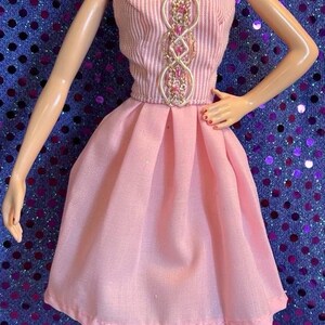 May include: A doll wearing a pink strapless dress with a pleated skirt and a decorative bodice. The doll has blonde hair, dangling earrings, and pink shoes. The background is a blue surface with purple sequins.