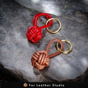 May include: Two leather keychains with knotted leather balls. One keychain is red and the other is brown. Both keychains have gold key rings.