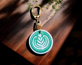 May include: A round, green and white keychain with a gold-colored clasp. The keychain features a latte art design with a heart and leaf pattern. The text "For Leather Studio" is at the bottom of the image.