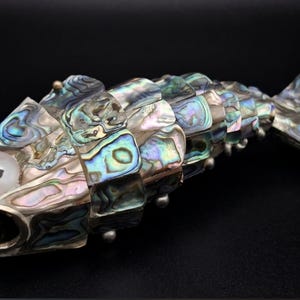 May include: A decorative fish figurine constructed from articulated segments inlaid with iridescent abalone shell. The fish features a prominent white circular eye, a wide open mouth, and a segmented body that displays a spectrum of blue, green, and pink hues.