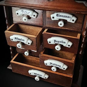 May include: A dark brown wooden spice cabinet with six drawers. Each drawer features a white ceramic handle plate labeled with black text: Cinnamon, Allspice, Paprika, Nutmeg, and Cloves. The cabinet has a decorative top edge and small knobs.
