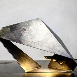 Może przedstawiać: Nowoczesna, geometryczna lampa stołowa wykonana z metalu o srebrnym wykończeniu. Lampa ma unikalny, kanciasty design i ciepłe, złote światło emanujące z podstawy.