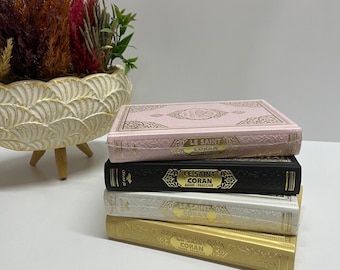 May include: A stack of four books with gold lettering and pink, black, white, and gold covers. The books are titled "Le Saint Coran" in French. The books are stacked on a white surface.