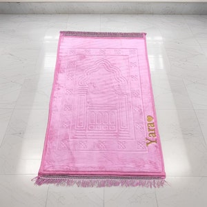 May include: A pink prayer rug with a traditional design and a gold embroidered heart with the name "Yara" on the side.