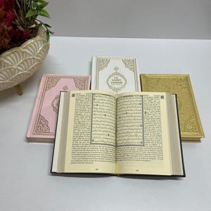 K&ouml;nnte beinhalten: Ein offener Koran mit schwarzem Einband und cremefarbenen Seiten. Der Koran ist auf einer Seite mit arabischem Text ge&ouml;ffnet. Im Hintergrund sind drei geschlossene Koranb&uuml;cher zu sehen, eines rosa, eines wei&szlig; mit goldenen Akzenten und eines golden.