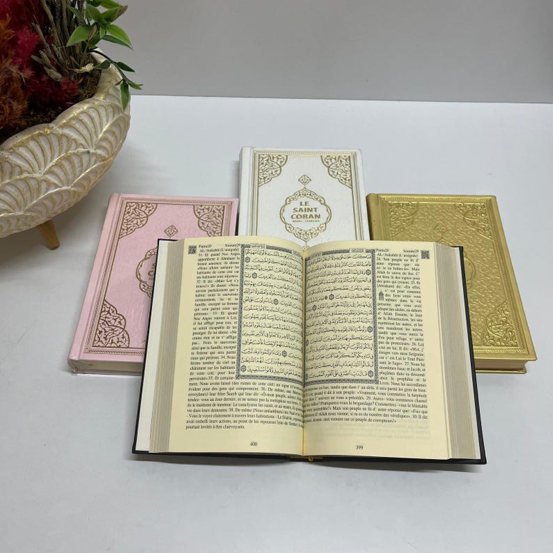 K&ouml;nnte beinhalten: Ein offener Koran mit schwarzem Einband und cremefarbenen Seiten. Der Koran ist auf einer Seite mit arabischem Text ge&ouml;ffnet. Im Hintergrund sind drei geschlossene Koranb&uuml;cher zu sehen, eines rosa, eines wei&szlig; mit goldenen Akzenten und eines golden.