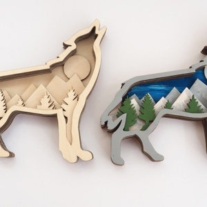 Könnte beinhalten: Zwei Wanddekorationen aus Holz in Form einer Wolfssilhouette mit einer mehrschichtigen Berg- und Waldszene im Inneren. Ein Wolf ist in natürlichen Holztönen bemalt, der andere in Grau mit blauen und weißen Akzenten.