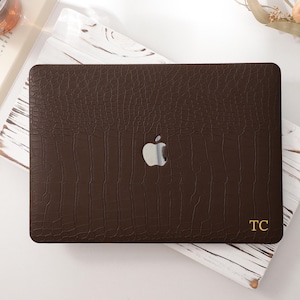 Può includere: Una custodia per laptop marrone scuro con una texture a pelle di coccodrillo. La custodia presenta un logo Apple argentato e le iniziali dorate "TC". La custodia è su una superficie di marmo bianco e grigio, con una penna, un libro e un bicchiere di liquido sullo sfondo.