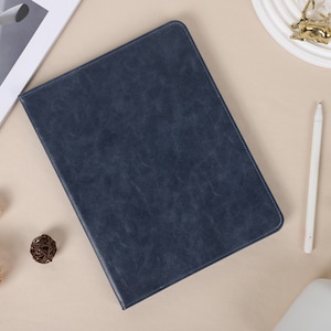 May include: A dark blue leather case for a tablet or e-reader. The case has a textured surface and is lying flat on a light brown surface.