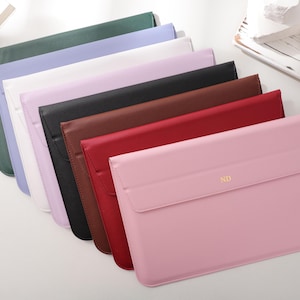 Op de afbeelding: Een verzameling laptophoezen in verschillende kleuren, waaronder groen, lavendel, wit, zwart, bruin, rood en roze. De roze hoes heeft de gouden initialen "ND". De hoezen zijn gemaakt van een glad materiaal en hebben een klepsluiting.