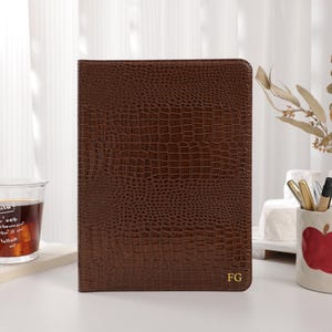 Peut inclure: Un journal en cuir marron avec une texture crocodile, personnalisé avec les initiales dorées "FG" en bas à droite. Le journal est posé sur une surface blanche, à côté d'un verre de thé glacé et d'un vase décoratif.