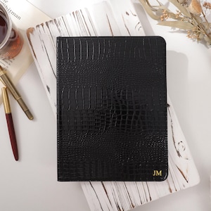 Puede incluir: Un diario de cuero negro con textura de piel de cocodrilo, con las iniciales doradas "JM" en la esquina. El diario descansa sobre una superficie de madera blanca, acompañado de un bolígrafo, un vaso de café y un libro.
