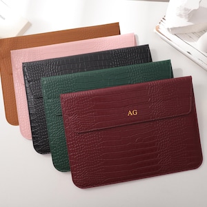 Peut inclure: Plusieurs pochettes pour ordinateur portable en cuir, de couleurs marron, rose, noir, vert et bordeaux. Chaque pochette présente un motif texturé façon crocodile. La pochette bordeaux porte les initiales dorées "AG".