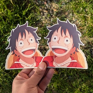 May include: Two die-cut stickers featuring a smiling cartoon character with black spiky hair, a red shirt, and a straw hat. The character has a large, open mouth and visible teeth. The stickers are held against a background of green grass.