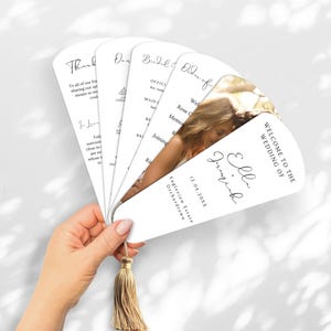 May include: White wedding fan with gold tassel, featuring text and a photo. The fan includes the words "Welcome to the Wedding of Ella & Jeremiah" and event details. The fan is held against a white background.