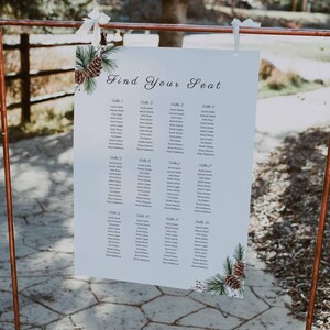 Puede incluir: Un plano de asientos de boda blanco con un tema rústico e invernal. El plano está impreso con las palabras "Find Your Seat" y enumera los nombres de los invitados por número de mesa. El plano se muestra en un soporte de cobre.