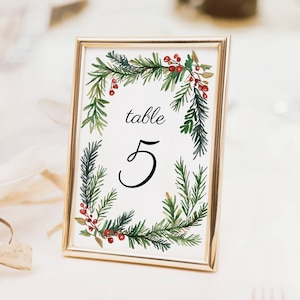 May include: A gold-framed table number card with a watercolor design. The card features the word "table" above the number "5" in a black script font. The number is surrounded by a wreath of green pine needles and red berries.