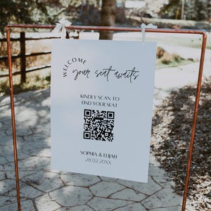 以下が含まれることがあります： 銅色の金属フレームが、エレガントな筆記体で「WELCOME your seat awaits」と書かれた白い看板を支えています。看板にはQRコードとSophia & Elijahの名前と日付も含まれています。看板は白いクリップで固定されています。