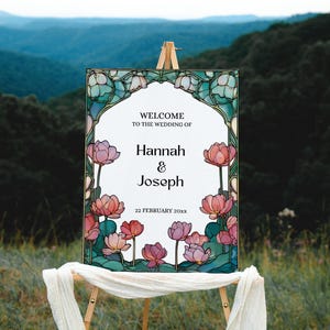 May include: A wedding welcome sign on an easel. The sign features a floral design with pink and red flowers and the names "Hannah & Joseph". The background is a scenic view of mountains and trees.