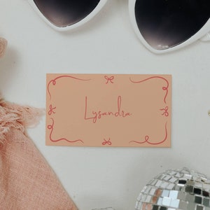 May include: A pink and white gift tag with a decorative border and the name "Lysandra" written in pink. The tag is sitting on a white surface with a pair of white heart-shaped sunglasses and a pink scarf.