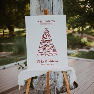 May include: A white wedding sign on a wooden easel. The sign has burgundy text: "Welcome to the Wedding of Holly & Nicholas 18 December 20XX." A burgundy tree design is in the centre.