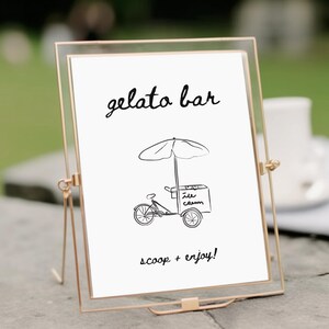 Könnte beinhalten: Ein goldgerahmtes Schild mit dem Schriftzug "gelato bar" in eleganter Schrift. Darunter eine skurrile Illustration eines Eiswagens mit Sonnenschirm. Der Text "scoop + enjoy!" vervollständigt das Design, ideal für eine Dessertausstellung.