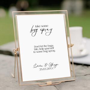 Puede incluir: Un marco rectangular de metal dorado que sostiene un letrero blanco con texto en negro que dice: take some bug spray, Don't let the bugs bite. help yourself to some bug spray. Emma & George 25.03.20XX. El marco tiene un soporte metálico fino.