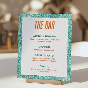 Puede incluir: Un letrero con marco de purpurina turquesa con las palabras "THE BAR" en naranja. El letrero enumera recetas de cócteles: "ACTUALLY ROMANTIC", "SHOWGIRL", "ELDEST DAUGHTER" y "OPALITE". El letrero está sujeto por un soporte de metal dorado.