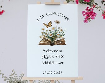 May include: A white sign with a floral illustration of an open book with a butterfly on top. The text reads "A New Chapter Begins" and "Welcome to Hannah's Bridal Shower 23.02.2025".