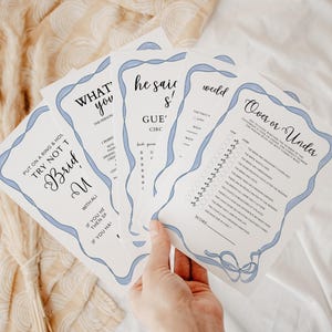 Op de afbeelding: Een set van vijf bruiloftsspelkaarten met blauwe lintaccenten. De kaarten bevatten spellen zoals "What's the Person You", "He Said She Said", "Over or Under" en "Try Not to Bride".