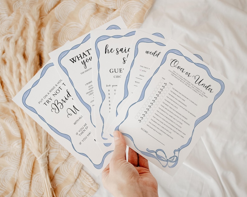 Op de afbeelding: Een set van vijf bruiloftsspelkaarten met blauwe lintaccenten. De kaarten bevatten spellen zoals "What's the Person You", "He Said She Said", "Over or Under" en "Try Not to Bride".